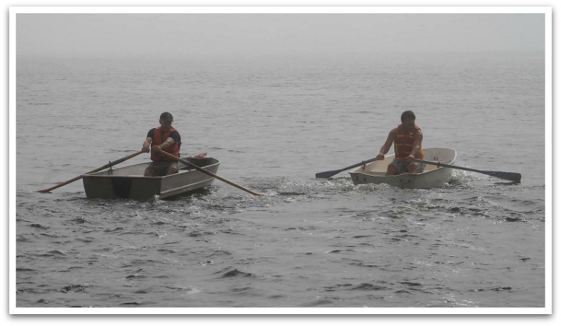 Two men racing each other in paddle boats on foggy waters.