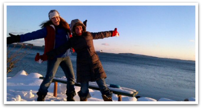 Two women in winter coats standing on snoe by a bench posing with their arms out and smiling. Ocean and island view in the background.